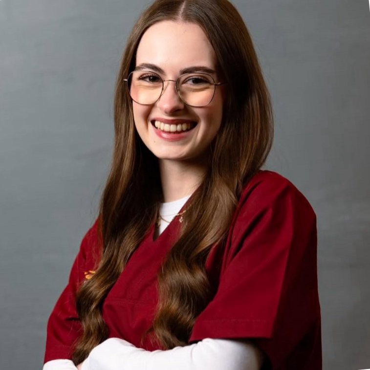 Person trägt ein weinrotes medizinisches oder pflegerisches Oberteil und steht vor einem grauen Hintergrund mit verschränkten Armen.