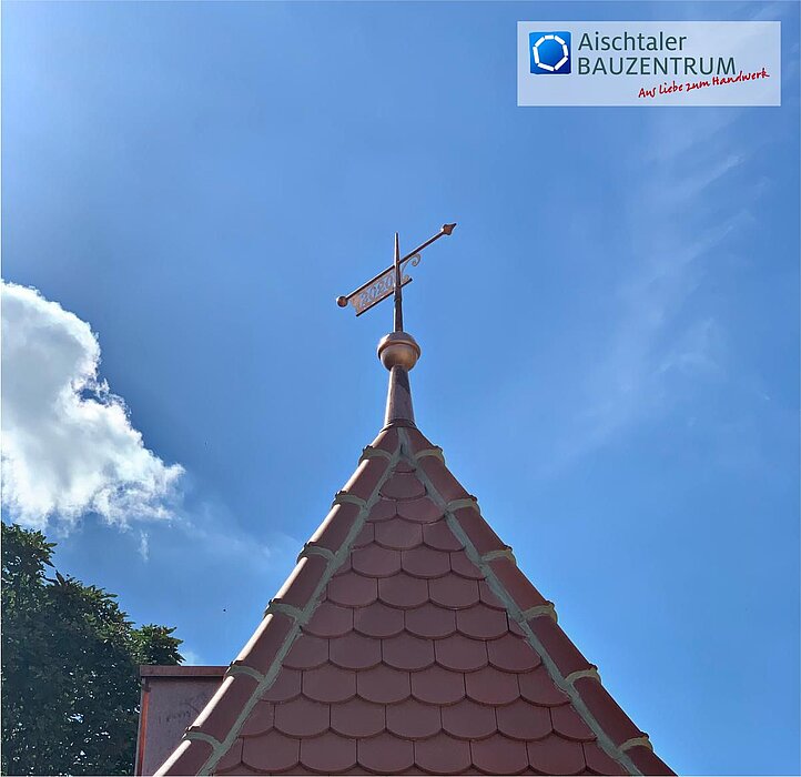 Spitzdach mit roten Dachziegeln und einer dekorativen Spitze, auf der eine metallene Wetterfahne mit der Aufschrift „NEA“ angebracht ist; im Hintergrund blauer Himmel mit einigen weißen Wolken und links unten grüne Baumkronen; oben rechts das Logo „Aischtaler Bauzentrum – Aus Liebe zum Handwerk“.