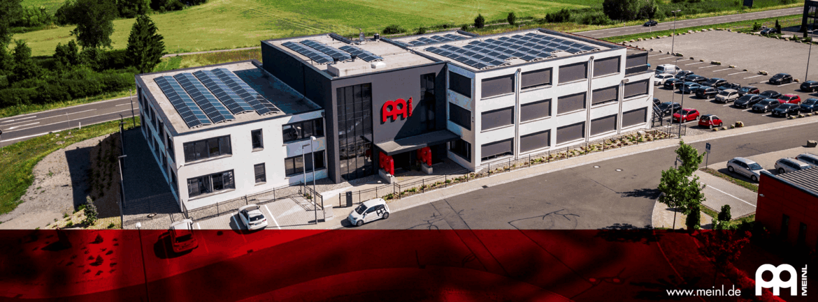 Ein modernes Firmengebäude mit grauer Fassade und großen Solarmodulen auf dem Dach steht an einer Straße, davor befinden sich mehrere geparkte Autos und ein Eingang mit rotem Akzent, auf der Front ist das rote Logo „Meinl“ zu sehen, unten rechts steht die Website „www.meinl.de“.