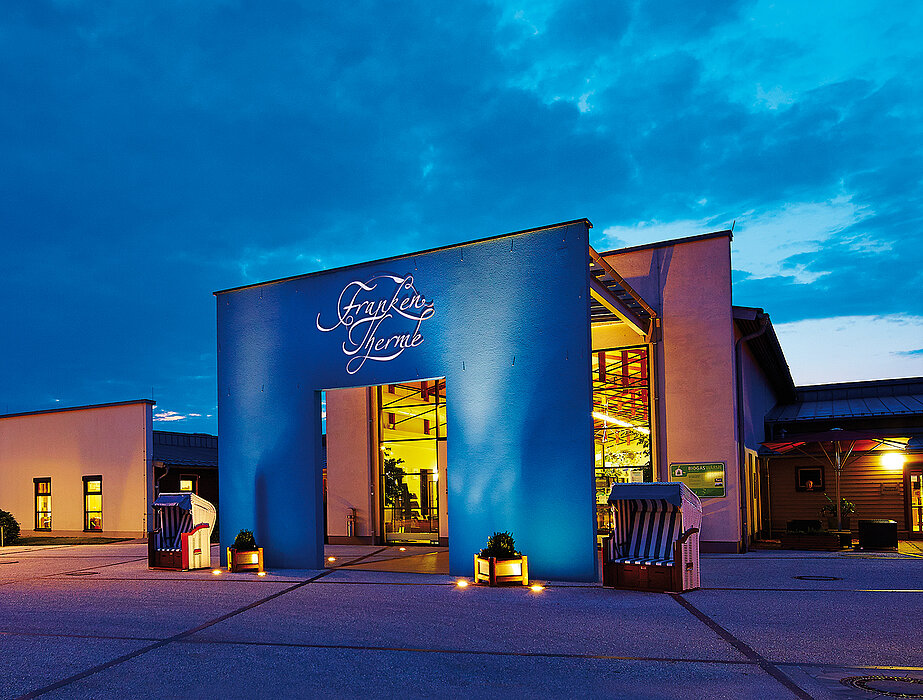 Ein beleuchteter Eingangsbereich der FrankenTherme mit blauem Portal und weißem Schriftzug „FrankenTherme“ steht vor einem modernen Gebäude, umgeben von Strandkörben und Pflanzen, unter einem dunkelblauen Abendhimmel.