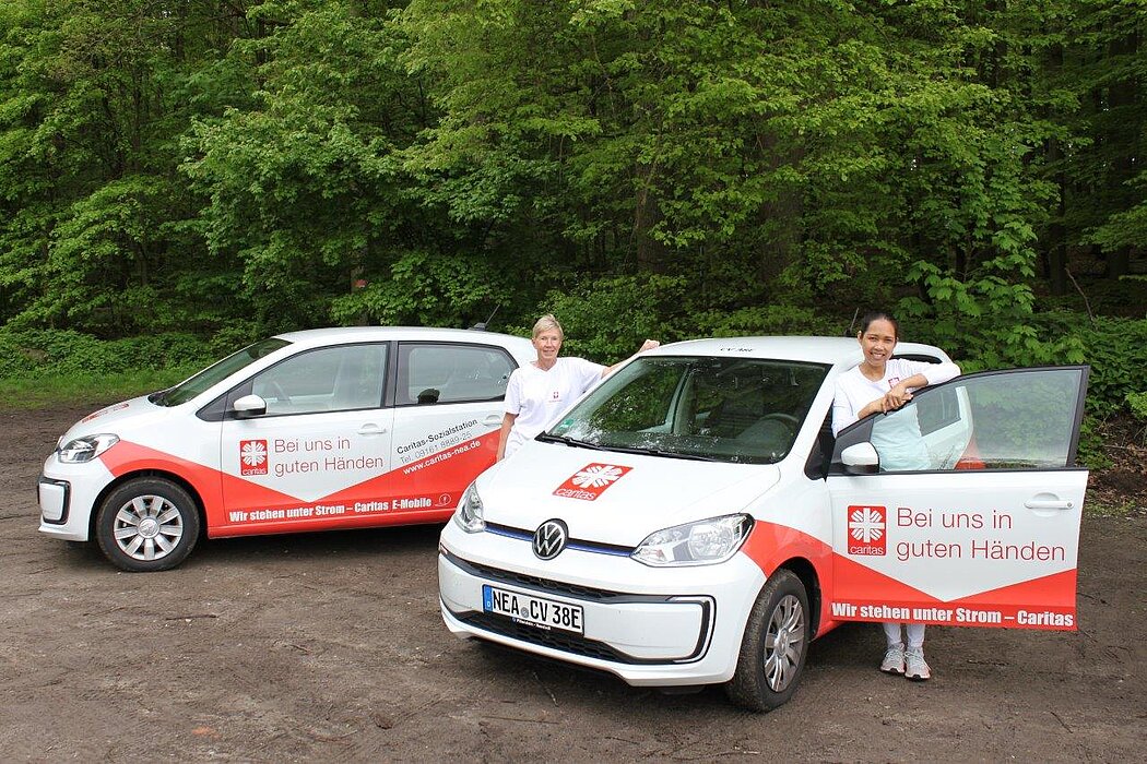 Zwei weiße Autos mit Caritas-Beschriftung und rotem Design, Text „Bei uns in guten Händen“ und „Wir stehen unter Strom – Caritas“, beide Fahrzeuge stehen auf einem Waldparkplatz, jeweils mit geöffneten Türen und Personen daneben.