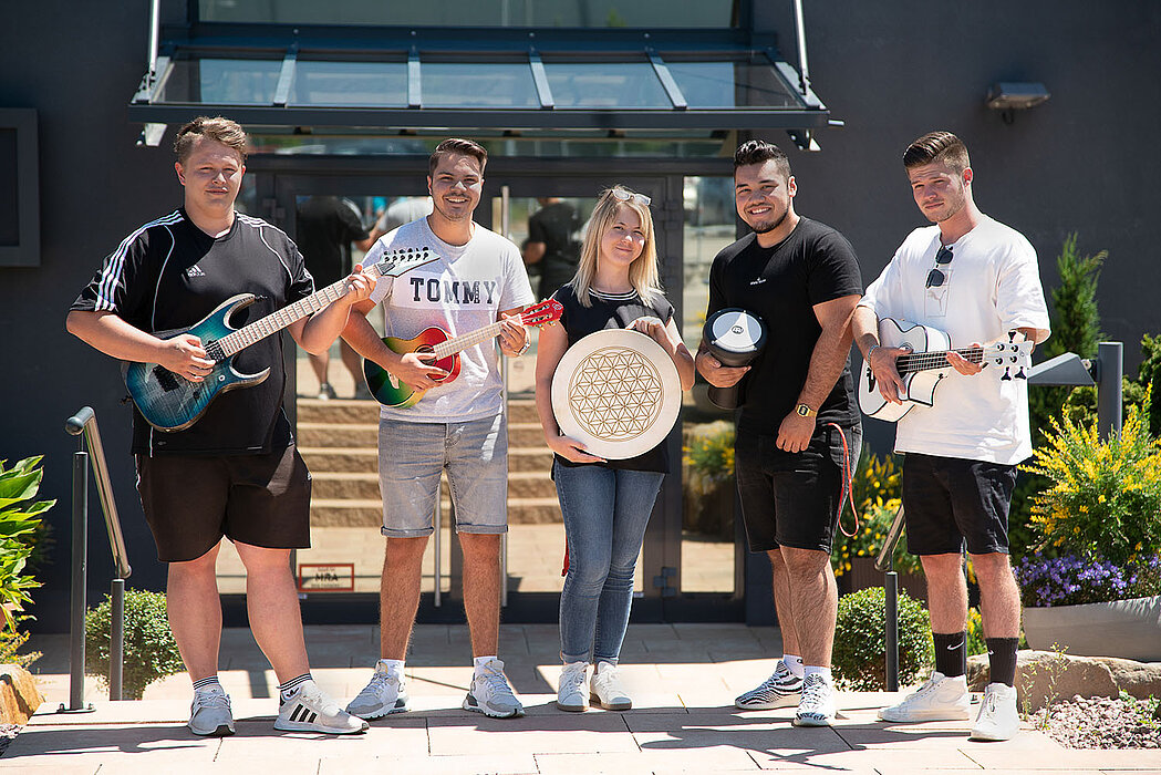 Fünf Personen stehen vor einem Gebäude und halten verschiedene Musikinstrumente, darunter zwei Gitarren, eine kleine rote Ukulele, eine Trommel und ein Megafon.