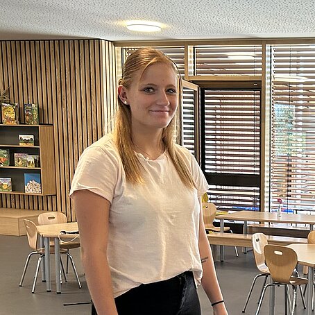 Zwei Personen stehen in einem hellen Raum mit großen Fenstern, modernen Möbeln und schlichter Einrichtung; sie tragen dunkle Kleidung und wirken in ein Gespräch vertieft.