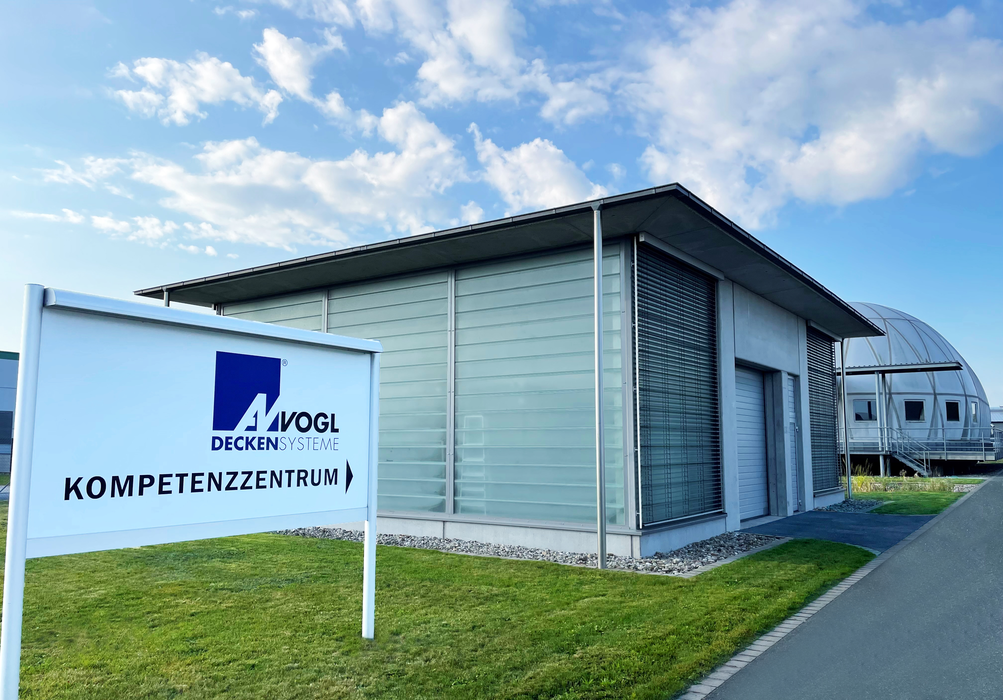 Gebäude mit moderner Glasfassade und grauen Lamellen, davor ein weißes Schild mit lila Logo und Text „Vogl Deckensysteme Kompetenzzentrum“, im Hintergrund eine Straße und ein rundes Gebäude mit Metallverkleidung unter blauem Himmel mit Wolken.