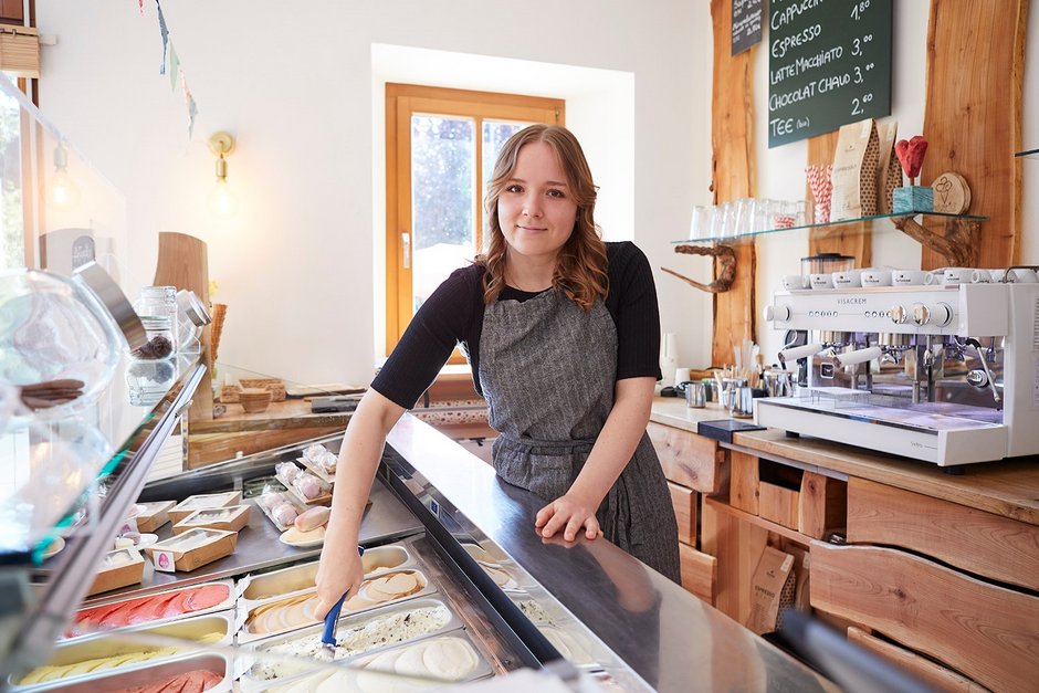 Person hinter Eistheke mit grauer Schürze schöpft Eis aus bunter Sortenauslage – rustikales Caféinterieur mit Espressomaschine, Menütafel und natürlichem Lichteinfall vermittelt warme, einladende Atmosphäre.
