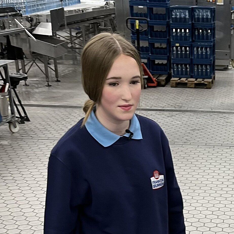 Eine Person in dunkelblauer HARIBO-Arbeitskleidung mit Headset steht in einer sauberen Produktionshalle mit Maschinen, Förderbändern und blauen Behältern im Hintergrund.