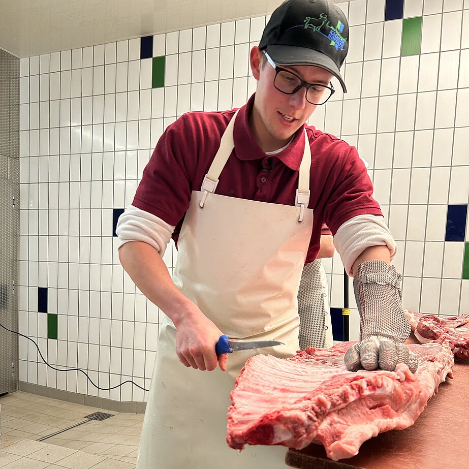 Eine Person mit weißer Schürze, rotem Shirt und Schutzkleidung zerteilt ein großes Stück rohes Fleisch in einer hellen, gekachelten Metzgerei oder Fleischverarbeitungseinrichtung.
