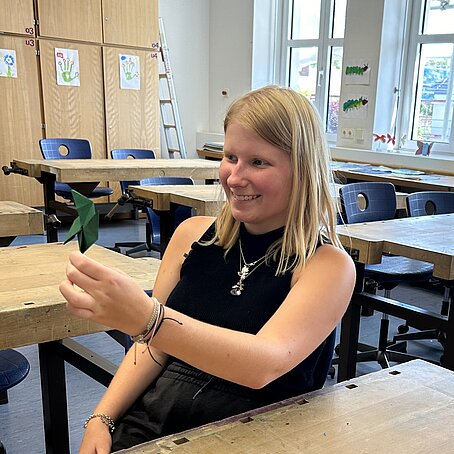 Ein Mädchen sitzt lächelnd in einem Klassenraum mit Holzmöbeln und blauen Stühlen, hält ein grünes Papier in der Hand; auf dem Tisch liegen weitere Bastelmaterialien, im Hintergrund sind Fenster, Zeichnungen und ein Regal mit Leiter sichtbar.
