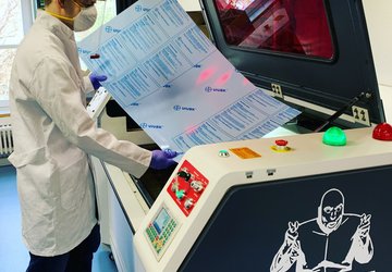 Mensch mit Schutzmaske und Schutzkittel steht an einem Lasercutter.