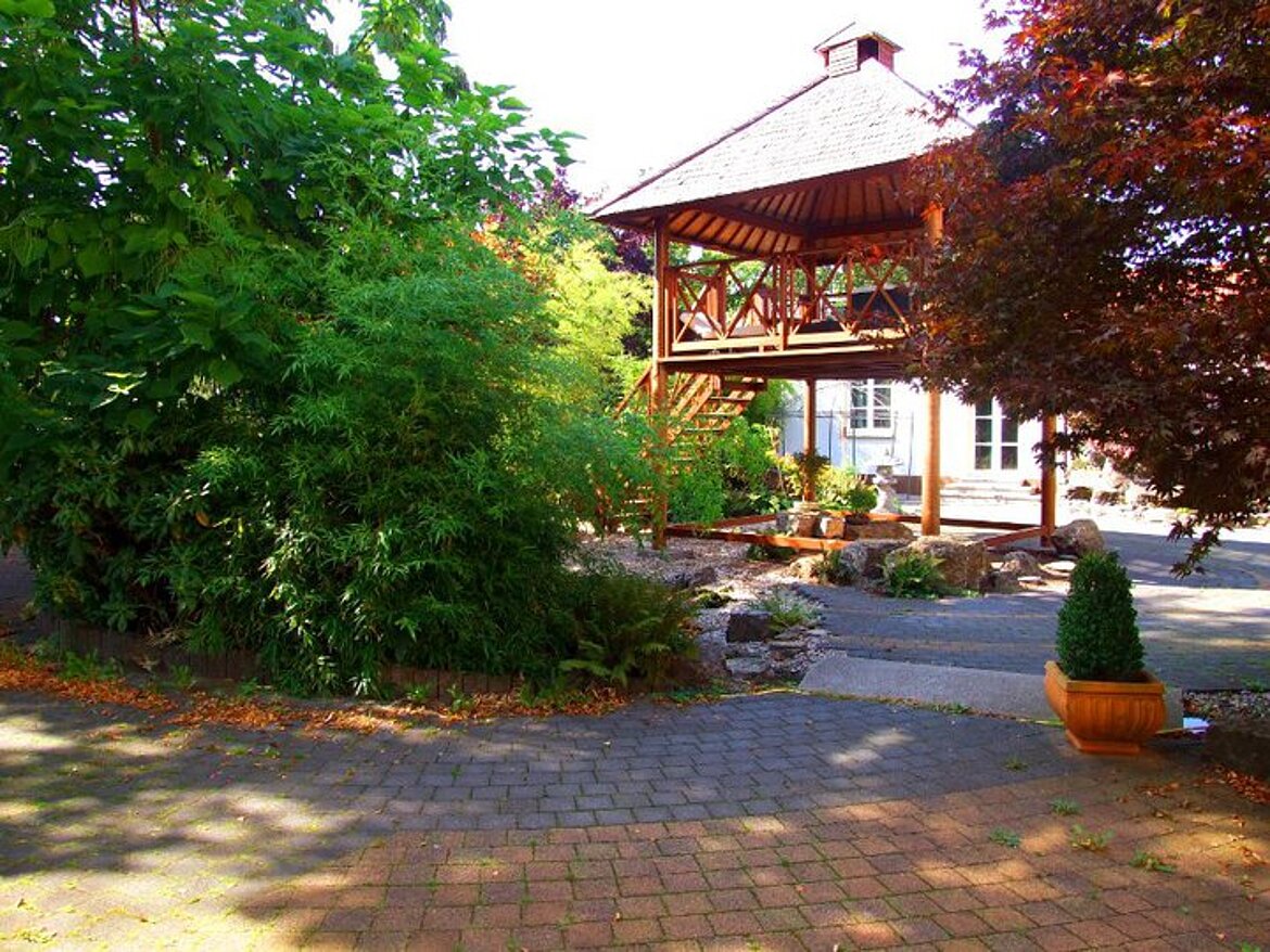 Ein überdachter Holzpavillon mit Treppe steht in einem Gartenbereich mit dichtem Grün, Kiesflächen, Pflanzkübeln und gepflastertem Weg.