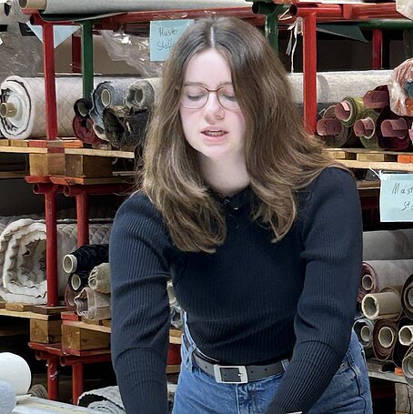 Eine Person mit langen braunen Haaren und Brille betrachtet an einem Tisch mehrere Druckmuster und Farbfelder; im Hintergrund befinden sich Regale mit aufgerollten Textilien in einem kreativ wirkenden Arbeitsraum.