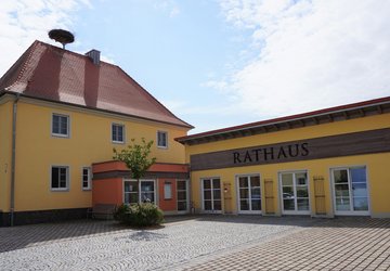 Zwei Gebäude auf einem gepflasterten Platz: links ein zweistöckiges Haus mit gelber Fassade, rotem Ziegeldach und einem Storchennest auf dem Dach; rechts ein eingeschossiges Gebäude mit der Aufschrift „RATHAUS“. Beide Gebäude haben Eingangsbereiche mit farblichen Akzenten. Vor dem Rathaus stehen Bänke und ein kleiner Baum.