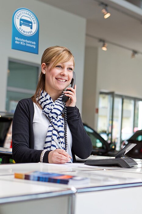 Person steht an einem Tresen, hält einen Telefonhörer und trägt einen weiß-blau gestreiften Schal, im Hintergrund ein Schild mit der Aufschrift „Meisterbetrieb der Kfz-Innung“.