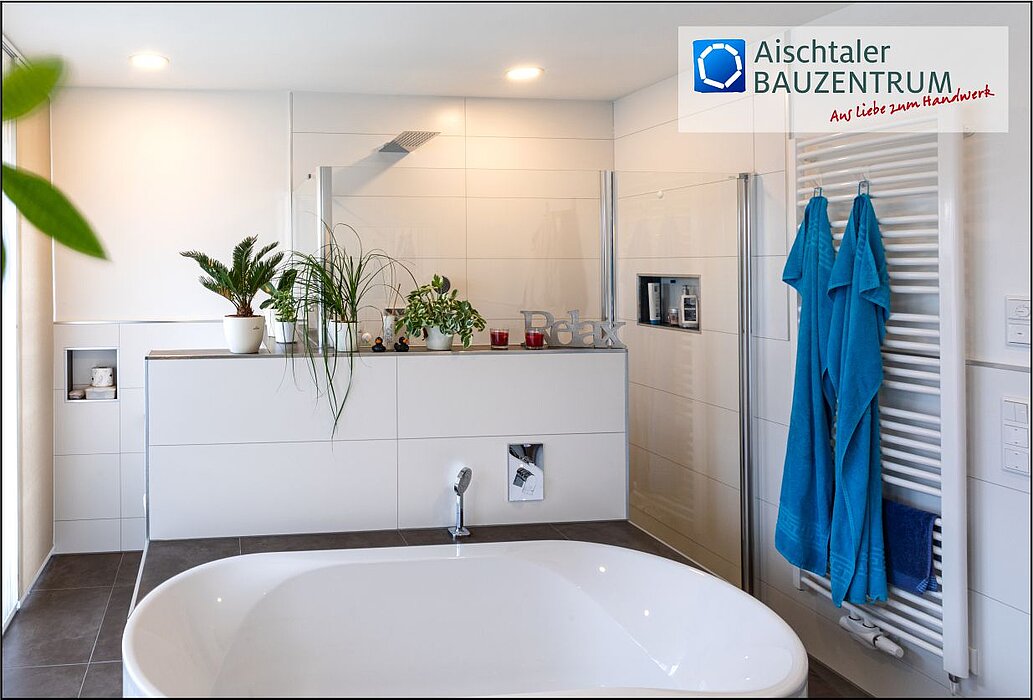 Modern Badezimmer mit freistehender weißer Badewanne im Vordergrund, dahinter eine weiße Wand mit eingelassenen Regalen, mehreren grünen Zimmerpflanzen und dekorativen Kerzen; rechts eine Glasdusche mit Regendusche und daneben ein weißer Handtuchheizkörper mit zwei blauen Handtüchern; Boden in dunklem Grau, helle Deckenbeleuchtung und oben rechts das Logo „Aischtaler Bauzentrum – Aus Liebe zum Handwerk“.