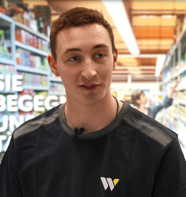 Eine Person trägt ein schwarzes Oberteil mit grauen Schulterpartien und einem weißen und gelben „W“-Logo, im Hintergrund sind Regale mit Produkten in einem Supermarkt.
